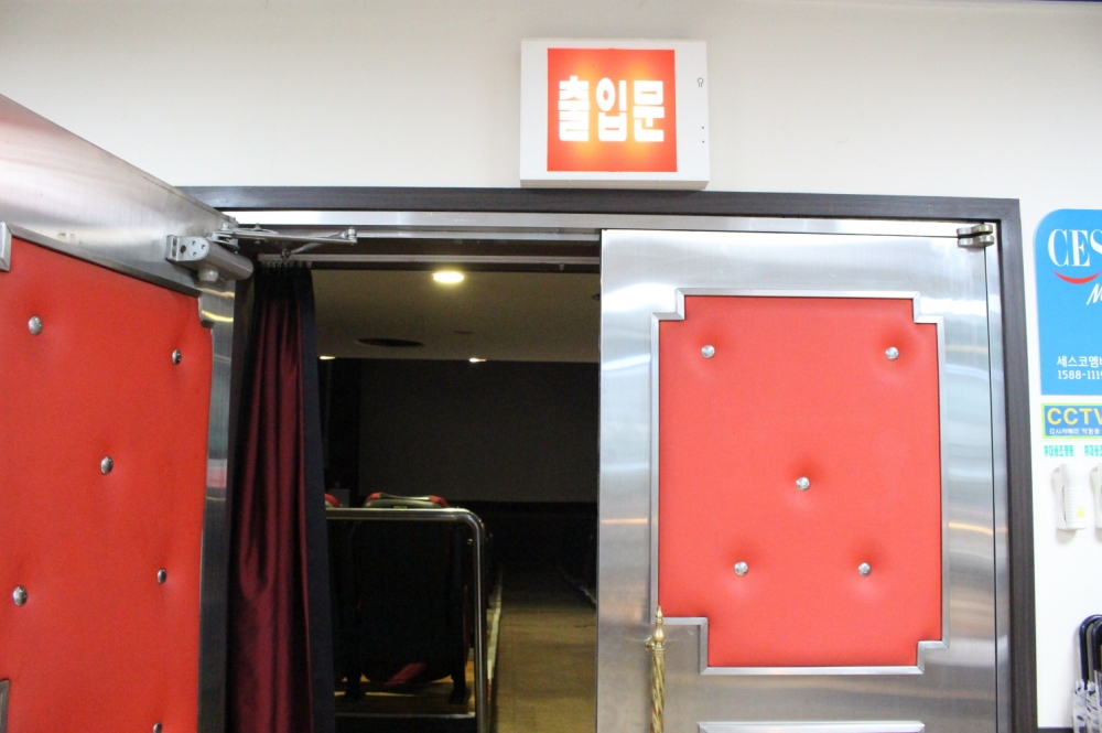 사진 설명 시작. 빨간색과 은색으로 이뤄진 두 문으로 구성된 상영관의 문이다. 왼쪽 문이 열려있고 그 사이로 객석이 보인다. 문 위에는 빨강 바탕에 흰 글씨로