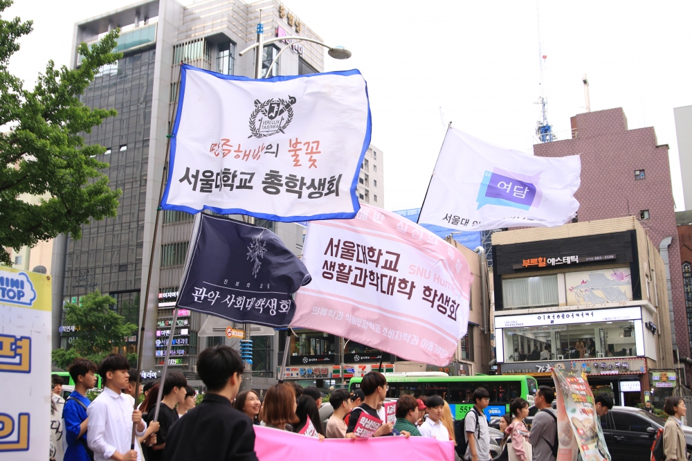 사진 설명 시작. 서울대입구역 주변 거리를 학생들이 행진하고 있다. 학생들이 들고 있는 깃발에는