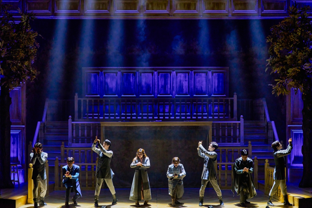 사진 설명 시작. 뮤지컬 「호프」의 2023년 공연 사진이다. 법정 사람들이 호프에 대해 수군대고 있다. 사람들은 법정을 배경으로 손으로 무언가를 적는 시늉을 하며 각자 앉고 서는 자세를 번갈아 가며 취하고 있다. 무대의 조명은 차가운 청색이고 인물들을 비추는 빛은 연한 노란빛이다. 사진 설명 끝.