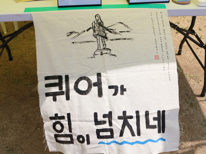 사진 설명 시작. 흰색 옥양천에 먹으로 퀴어가 힘이 넘치네 라는 문구가 크고 동글동글하게 쓰여 있다. 문구 위에는 종아리까지 오는 짧은 한복치마를 입고 댕기머리를 드리운 소양강 처녀상이 산을 배경으로 우뚝 서 있는 모습이 먹으로 그려져 있다. 사진 설명 끝.