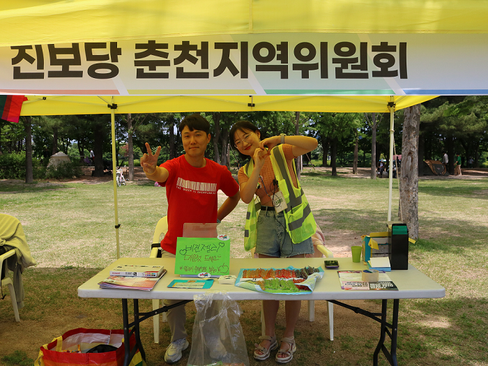 사진 설명 시작. 진보당 춘천지역위원회 라고 쓰인 부스 아래 젊은 남성과 노란 스탭 조끼를 입은 젊은 여성이 포즈를 취하고 있다. 남성은 손가락으로 브이자를 그리고 있고, 여성은 두 손으로 하트를 만들고 있다. 그들 앞 탁상에는 비건젤리가 진열돼 있으며, 옆에는 비건젤리 마음껏 드세요 라는 문구가 쓰여 있다. 사진 설명 끝.