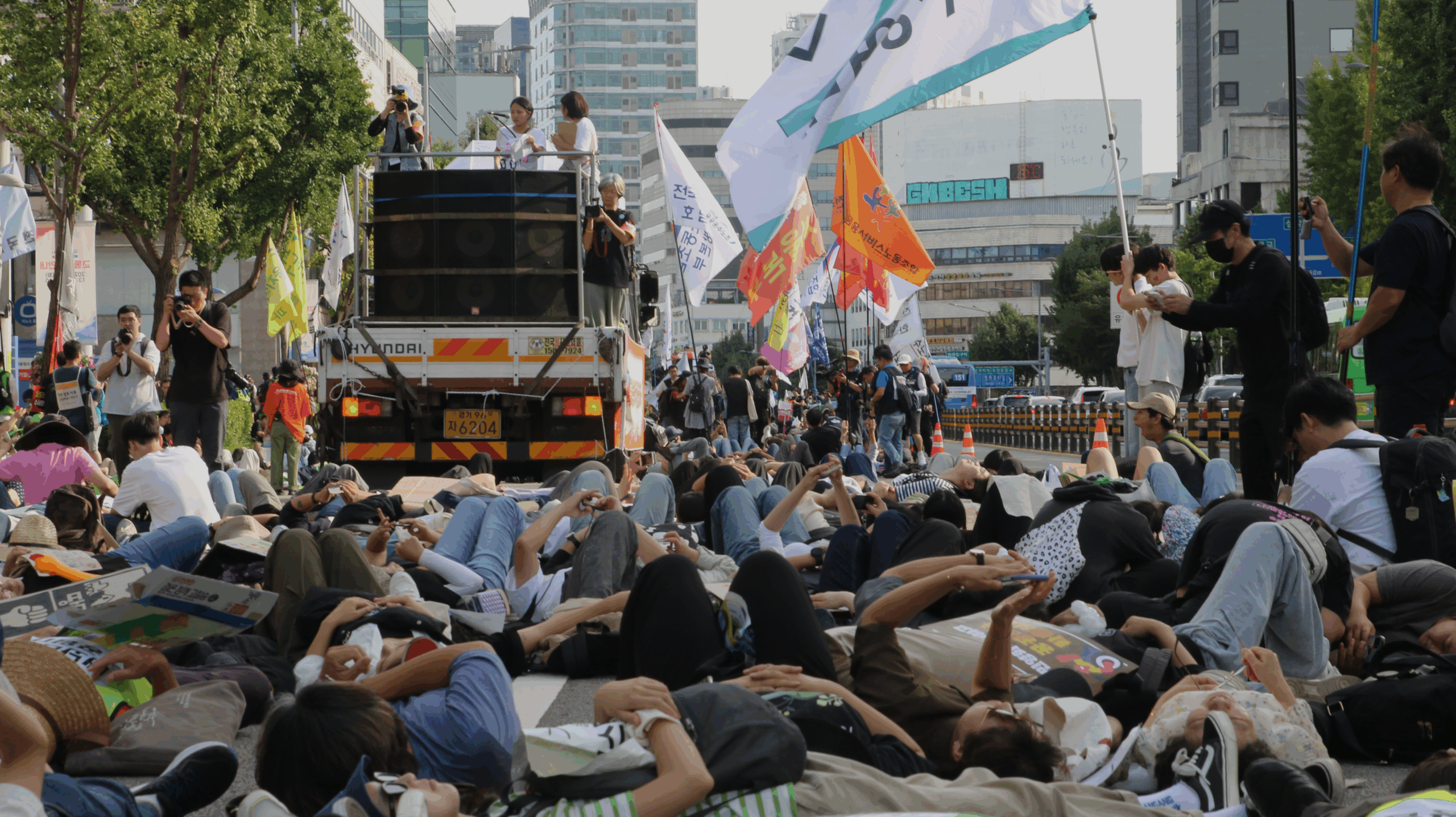 사진 설명 시작. 사람들이 다이인 퍼포먼스를 위해 모두 길 한복판에 누워 있다. 화면 좌측에는 스피커가 장착된 차량에서 위험을 알리는 경보 사이렌이 나오고 있다. 사진 설명 끝.