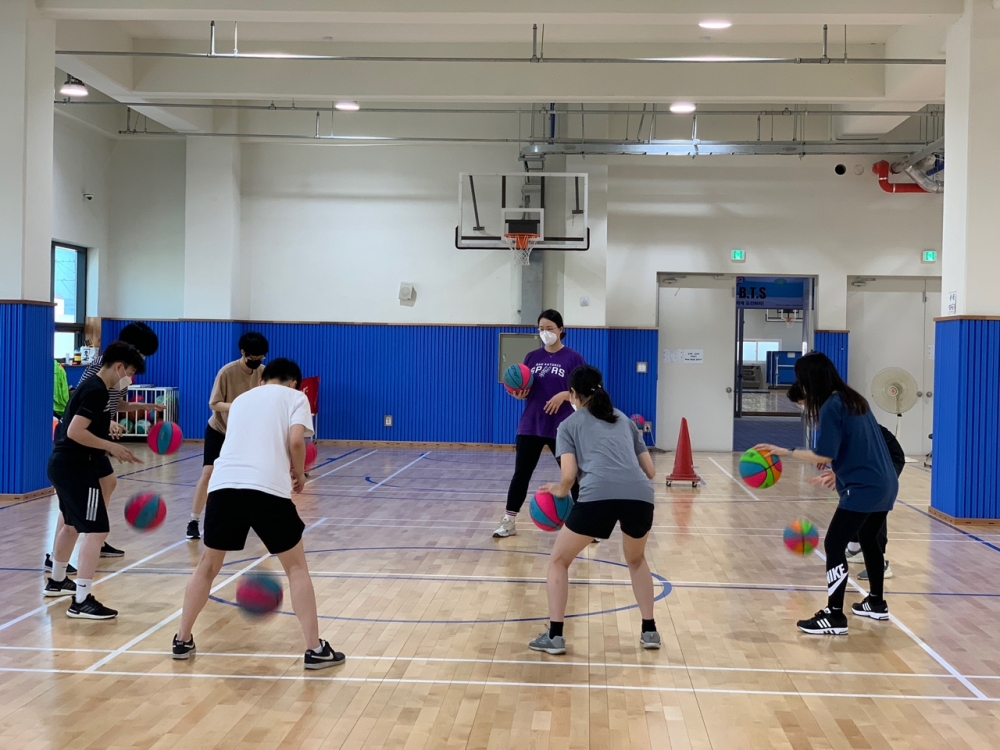 사진 설명 시작. 실내 체육관의 농구 코트에서 여성들이 농구공으로 연습을 하고 있다. 가장 뒤에 농구골대가 있으며 그 앞에 강사로 보이는 여성이 공을 들고 서 있다. 나머지 여성 여섯 명은 손바닥으로 농구공을 튕기는 동작을 하고 있다. 사진 설명 끝.