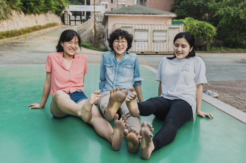 사진 설명 시작. 변화의월담 운영진 세 명이 농구장에 다리를 뻗고 나란히 앉아 있다. 맨발로 한창 운동한 뒤라 발바닥이 새카맣다. 한쪽 발을 들며 환하게 웃고 있다. 사진 설명 끝