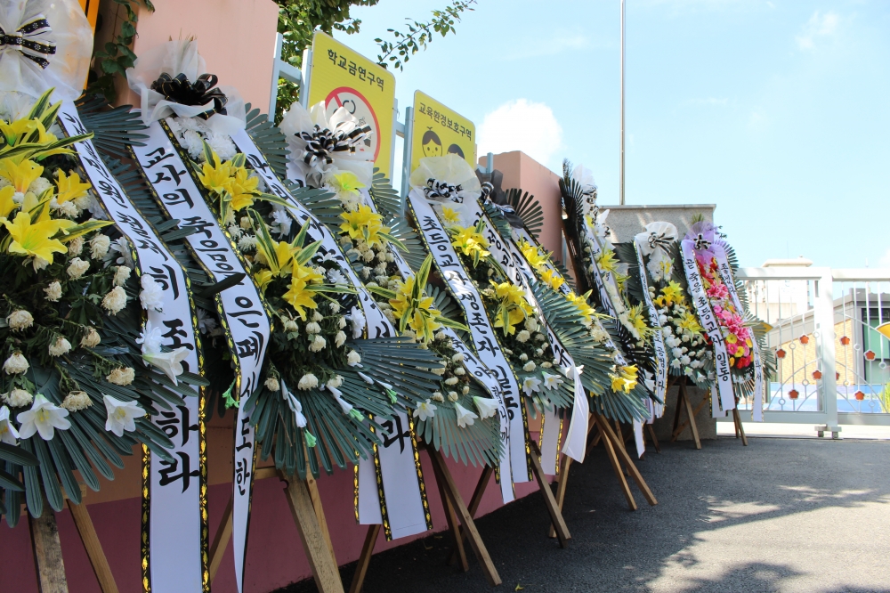 사진 설명 시작. 파란 하늘 교문 아래 근조화환들이 늘어서 있다. 화환에는