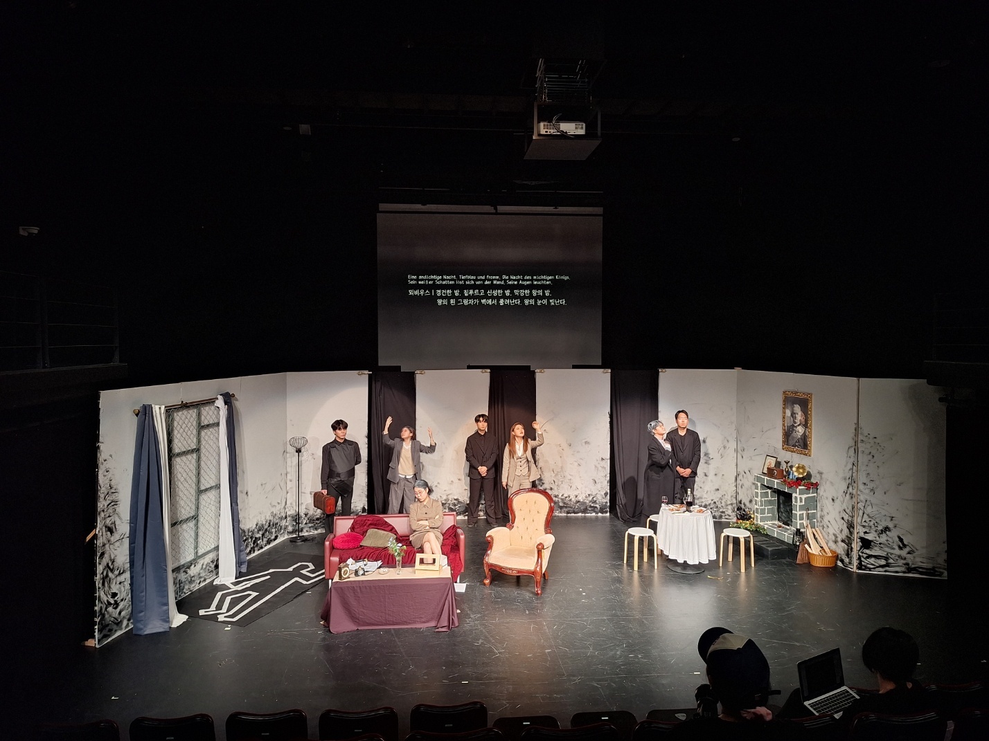 사진 설명 시작. 연극이 진행되는 무대. 무대에는 소파와 탁자가 놓여 있고, 그 사이에서 일곱 명의 배우들이 연기를 하고 있다. 사진 설명 끝.