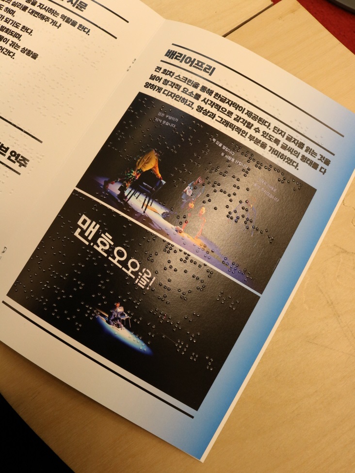 사진 설명 시작. 연극 견고딕 걸의 프로그램북 중 배리어프리 요소들을 설명하는 페이지다. 페이지 위에는 배리어프리라는 단어가 볼드체로 쓰여 있고, 아래에는 부연설명과 공연 사진 두 장이 삽입돼 있다. 부연설명은 다음과 같다. 전 회차 스크린을 통해 한글자막이 제공된다. 단지 글자를 읽는 것을 넘어 청각적 요소를 시각적으로 감각할 수 있도록 글씨의 형태를 다양하게 디자인하고, 영상과 그래픽적인 부분을 가미하였다. 사진 설명 끝.