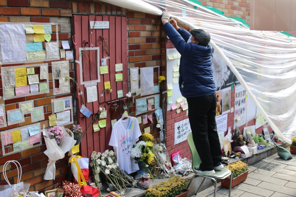 사진 설명 시작. 추모 메시지와 꽃다발이 비에 젖지 않도록 비닐로 감싸주는 시민의 모습이 보인다. 사진 설명 끝.