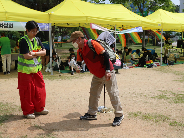 사진 설명 시작. 중년 남성이 제기차기를 위해 제기를 던지는 움직임을 취하고 있다. 그 모습을 행사 스태프가 지켜보고 있다. 뒤의 노란 천막에 사람들이 옹기종기 모여 앉아있다. 그들의 머리 위에 무지개빛 천들이 펄럭인다. 사진 설명 끝.