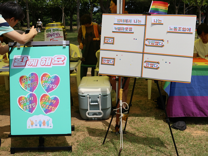 사진 설명 시작. 왼쪽에는 대한성공회 춘천나눔의집 부스의 이벤트 안내 판넬이, 오른쪽에는 민주노총 강원지부의 이벤트 판넬이 세워져 있다. 왼쪽 판넬에는 다음의 문구가 쓰여 있다. 성소수자 공부모임, 성소수자 신앙상담, 성소수자 자조모임 함께 해요. 오른쪽 판넬에는 다음의 문구가 쓰여 있다. 일터에서 나는 커밍아웃을 했다, 나는 노동조합에 가입했다. 사진 설명 끝.