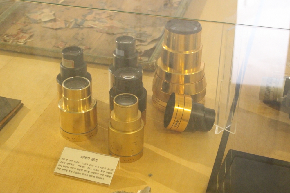 사진 설명 시작. 다양한 종류의 카메라 렌즈가 6점 전시돼 있다. 렌즈는 금색과 검정색이 섞였고 다양한 크기다. 가장 큰 것은
