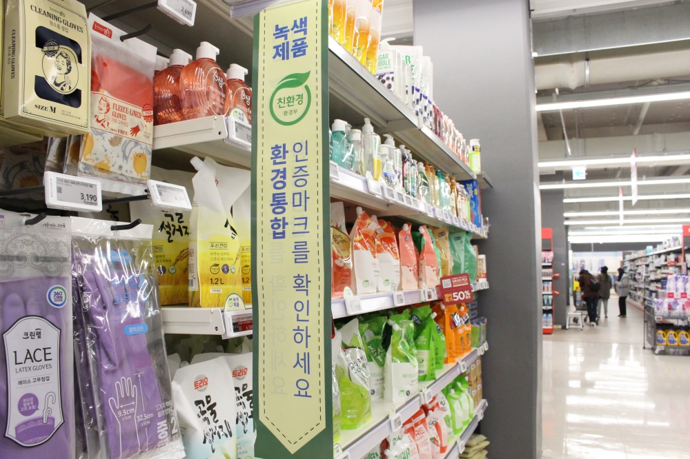 사진 설명 시작. 가판대에 각양각색의 세제 제품들이 진열돼 있다. 가판대 위쪽에는 친환경 제품임을 알리는 초록색 배너가 달려 있다. 배너에는 파란 글씨로 녹색 제품, 환경종합 인증마크를 확인하세요 라는 글씨가 쓰여 있고, 중간에 환경부의 친환경 마크가 붙어 있다. 사진 설명 끝.