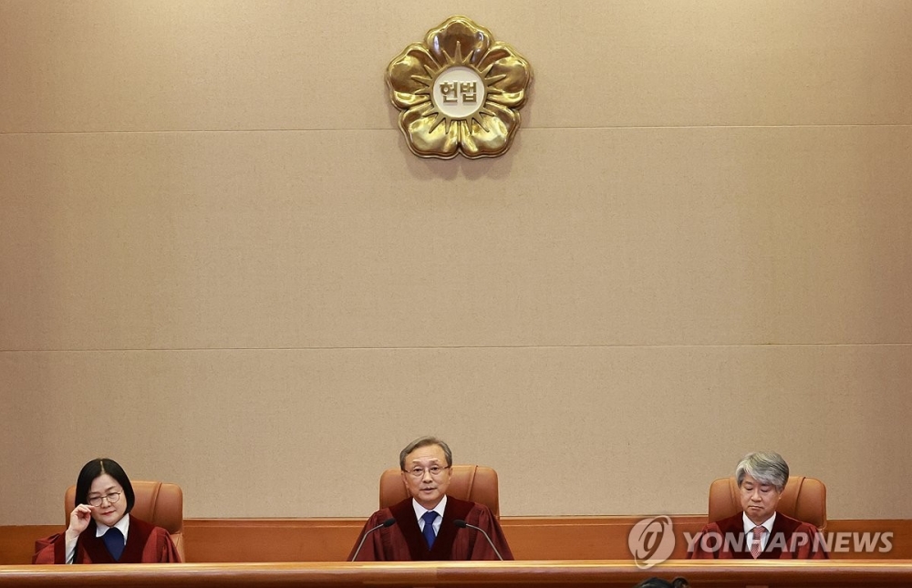 사진 설명 시작. 헌법재판소 사진이다. 무궁화의 외형 안에 ‘헌법’이라고 적힌 문양이 제일 위에 있고, 아래에 헌법재판관 세 명이 앉아서 선고하고 있는 모습이다. 우측 하단에는 ‘YONHAP NEWS’ 워터마크 로고가 있다. 사진 설명 끝.