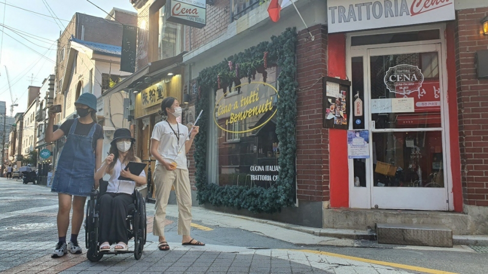 사진 설명 시작. 샤로수길 거리에 서배공 세 사람이 서있다. 오른쪽에는 가게가 있다. 세 사람 중 왼쪽 사람은 핸드폰으로 사진을 찍고 있다. 가운데 사람은 태블릿을 들고 휠체어를 타고 있다. 가장 오른쪽의 사람은 자를 들고 가게를 바라보고 있다. 사진 설명 끝.