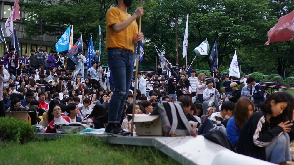 사진 설명 시작. 중앙도서관 앞 아크로폴리스에 학생들이 가득차 있다. 학생들은 다양한 단체의 깃발을 들고 있다. 사진 설명 끝.