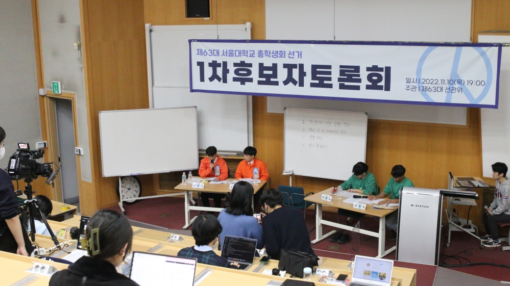 사진 설명 시작. 강의실 연단에 양 선본의 후보자들이 나란히 앉아 있다. 후보자들 위에