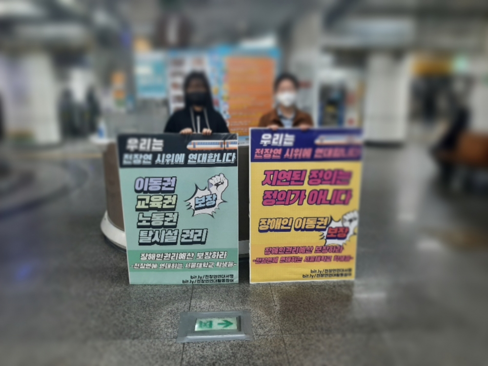 사진 설명 시작. 지하철 역사 내에서 두 사람이 몸집만한 피켓을 들고 나란히 서있다. 얼굴과 배경은 블러 처리됐다. 피켓에는 왼쪽부터