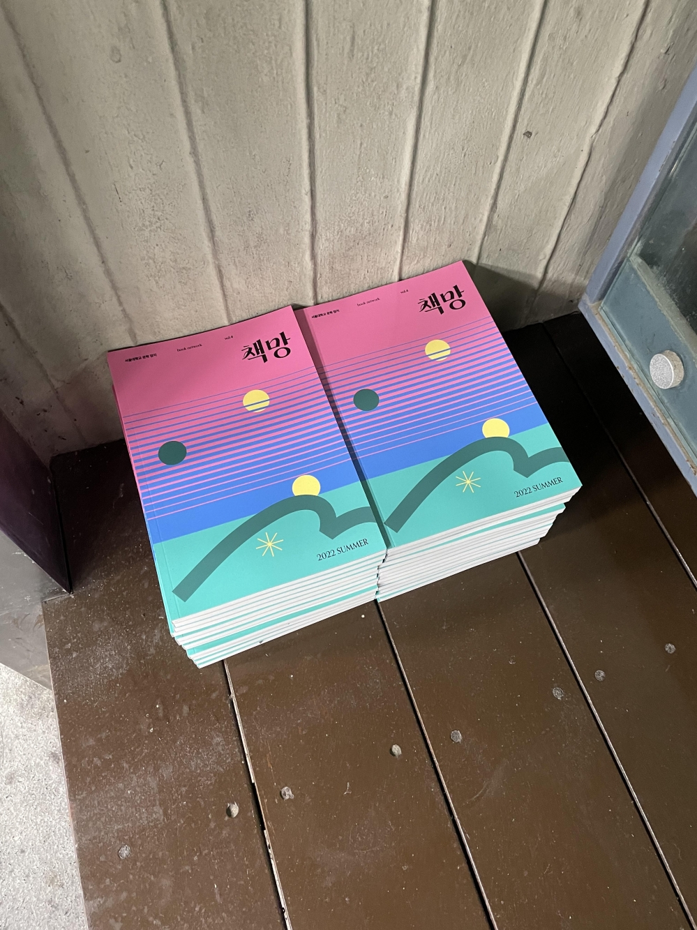 사진 설명 시작. 나무 판자 위에 여러 권의 책망 문예지가 쌓여있다. 책의 표지는 분홍, 파랑, 민트색이 그라데이션으로 칠해져 있고, 중앙 부근에 여러 개의 가로 선과 동그라미, 눈송이 모양, 곡선 등의 도형이 위치해있다. 책의 가장 아래 쪽에는