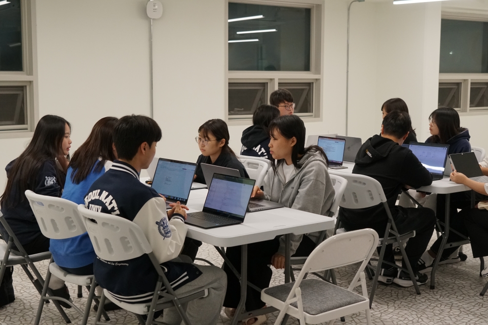 사진 설명 시작. 마주보게 배치된 흰색 탁자에 학생들이 모여앉아있다. 학생들은 각자의 노트북으로 자료를 찾아보고 있다. 사진 설명 끝.