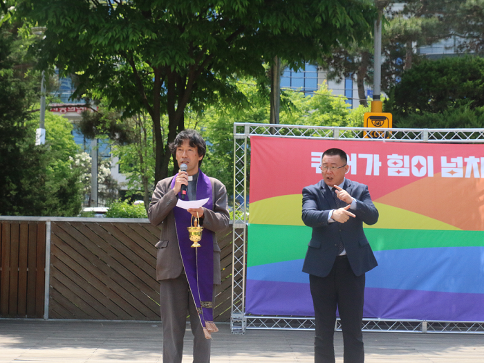사진 설명 시작. 퀴어가 힘이 넘치네 라는 문구가 쓰인 무지갯빛 현수막이 설치된 무대이다. 무대 앞에는 사제복을 입은 양만호 사제가 한 손에 향유를, 다른 손에 마이크를 들고 축복기도를 낭독하고 있다. 양만호 사제의 오른쪽에는 수어통역사가 기도내용을 통역하고 있다. 사진 설명 끝.