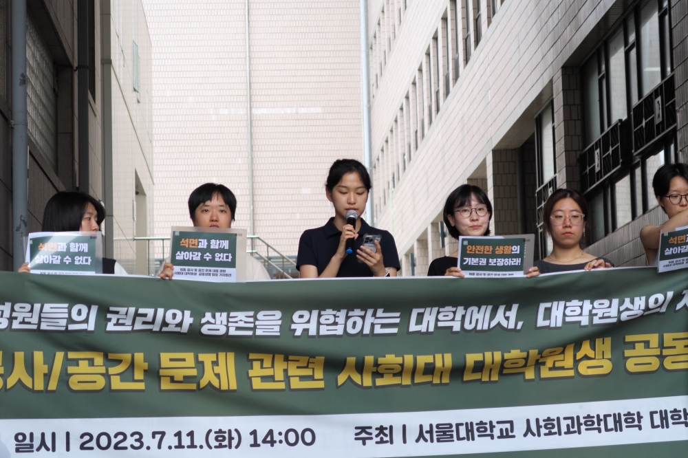 사진 설명 시작. 강다겸 씨가 마이크를 쥐고 발언하고 있다. 강 씨의 옆에는 다른 대학원생들이 피켓을 들고 서 있다. 사진 설명 끝.