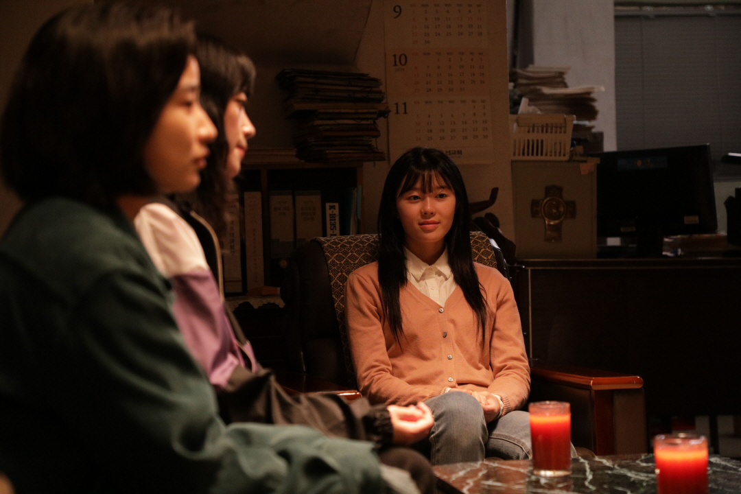 사진 설명 시작. 어두운 조명 아래, 선우, 나미, 채린이 의자에 앉아 있다. 세 인물은 정면을 보고 있다. 채린은 웃고 있고, 선우와 나미는 무표정이다. 사진 설명 끝.