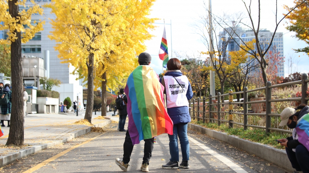 사진 설명 시작. 행진 도중 쉬는 시간 행진 참여자 두 명의 뒷모습. 왼쪽 사람은 커다란 무지갯빛 깃발을 숄처럼 몸에 두르고 있고, 오른쪽의 사람은 차별금지법 연내 제정하자라는 문구가 적힌 흰색 조끼를 입고 있다. 사진 설명 끝.