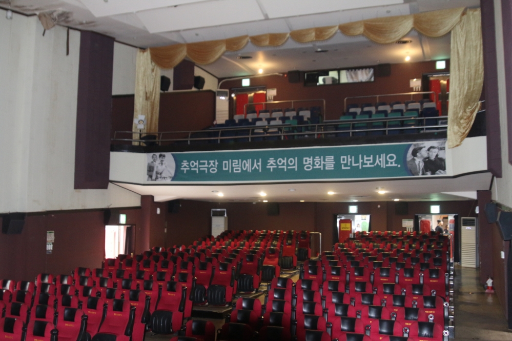 사진 설명 시작. 2층으로 구성된 영화상영관의 모습이다. 1층에는 빨간 의자가 2층에는 초록 의자, 회색 의자가 있다. 2층 위에는 상영실이 보인다. 2층의 덮개가 시작되는 돌출된 부분에