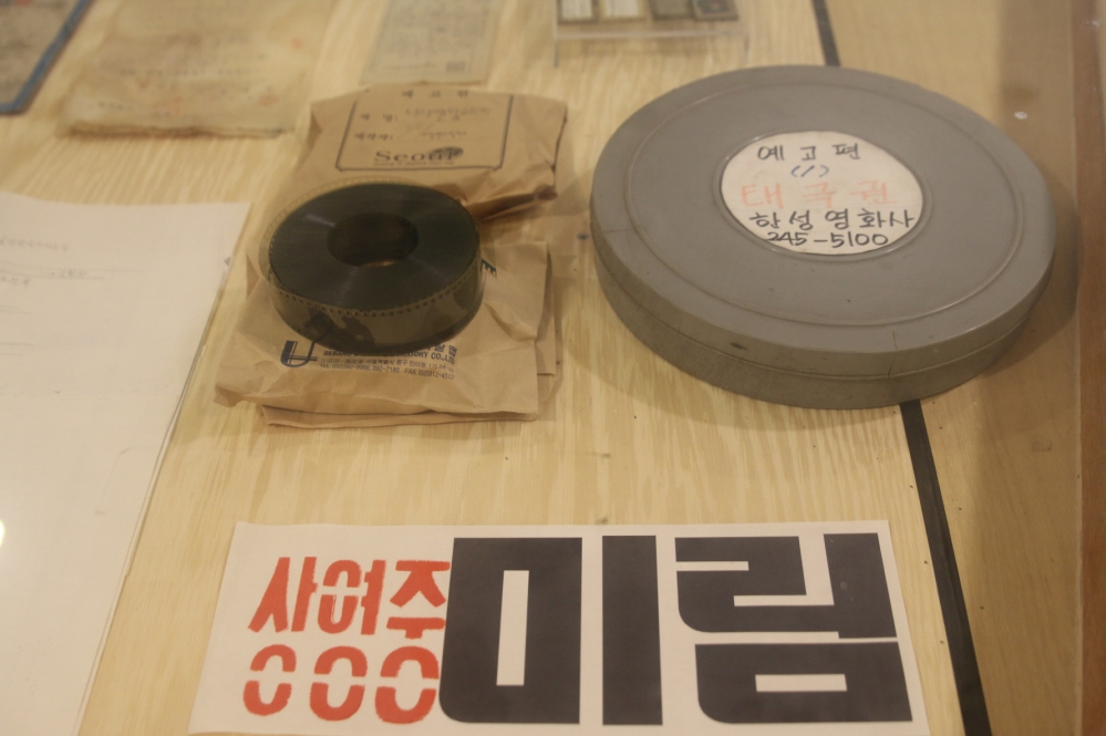 사진 설명 시작. 상단 오른쪽에 동그란 회색 필름통이, 왼쪽에 동그랗게 말린 검정 필름이 있다. 회색 필름통 위의 흰색의 동그란 공간에는