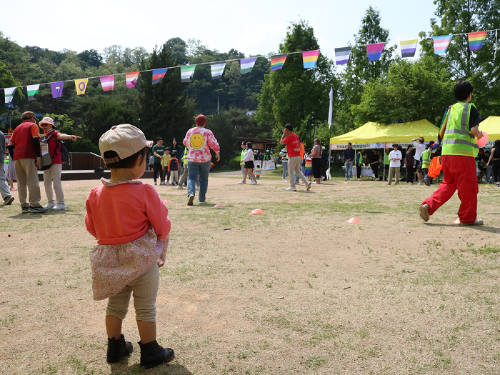 사진 설명 시작. 색색의 깃발 아래 참가자들이 삼삼오오 모여 있다. 가까이에 모자를 쓴 아이의 뒷모습이 보인다. 사진 설명 끝.