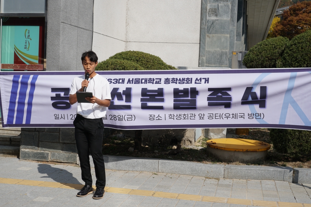 사진 설명 시작. 공명 부후보 정영훈 씨가 마이크와 태블릿을 든 채 발언하고 있다. 뒤로는 ‘제63대 서울대학교 총학생회 선거 공동선본발족식’이라는 글씨와 일시, 장소가 적힌 현수막이 가로로 걸려 있다. 사진 설명 끝.