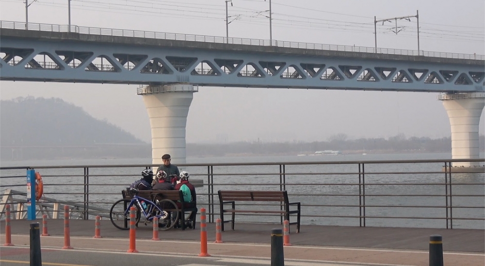 사진 설명 시작. 한강 다리 밑으로 흐르는 강이 보인다. 그 앞에 놓인 벤치에 헬멧을 쓴 세 명의 사람이 앉아 있다. 그들 뒤로는 자전거가 한 대 보인다. 사진 설명 끝.