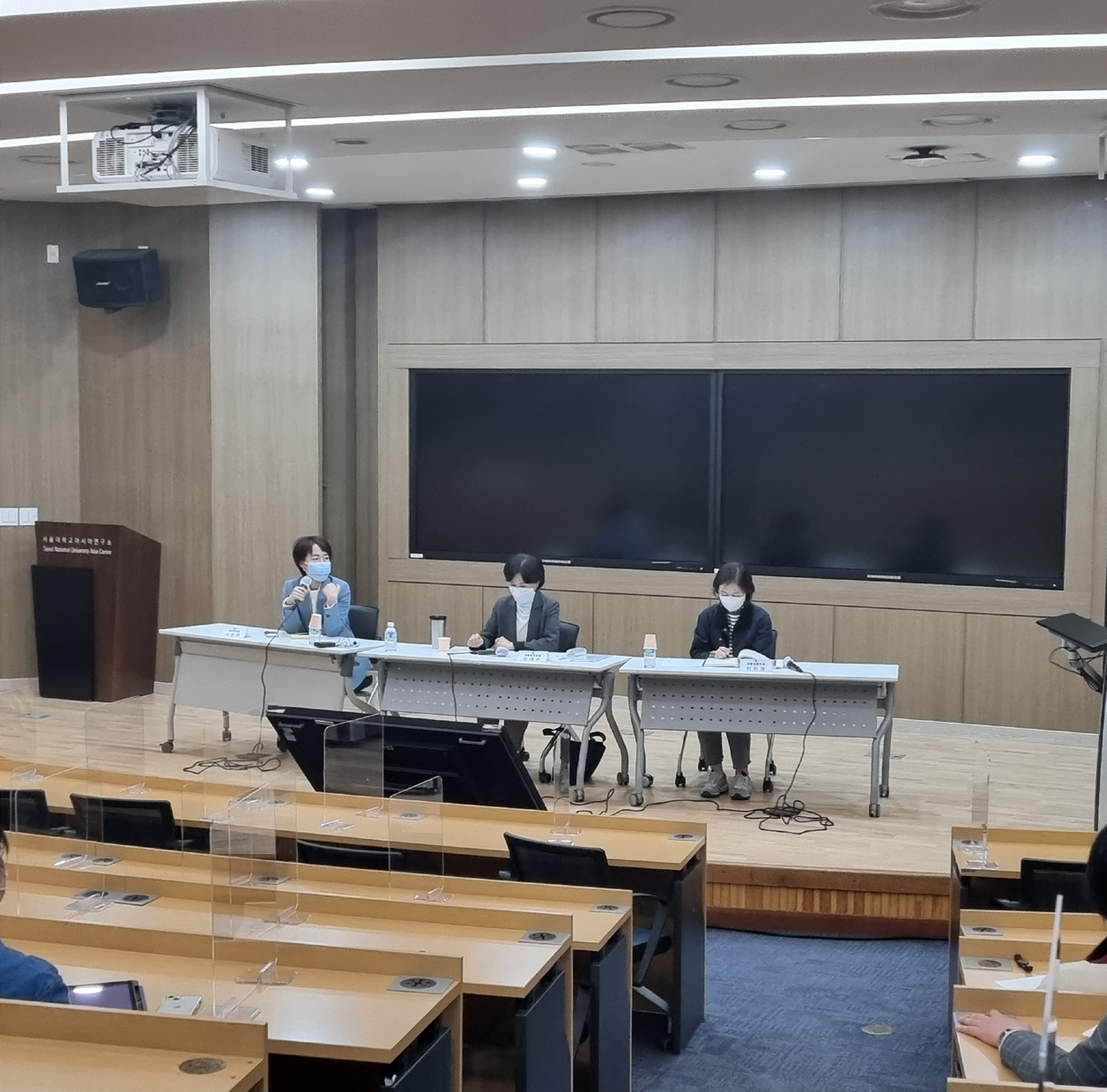 사진 설명 시작. 강의실 단상에 책상 세 개가 놓여있고 각 책상마다 사람이 한 명씩 앉아있다. 가장 좌측에 있는 중년 여성이 마이크를 들고 발언하고 있다. 강의실 좌석 쪽에는 사람들이 듬성듬성 앉아있다. 사진 설명 끝.