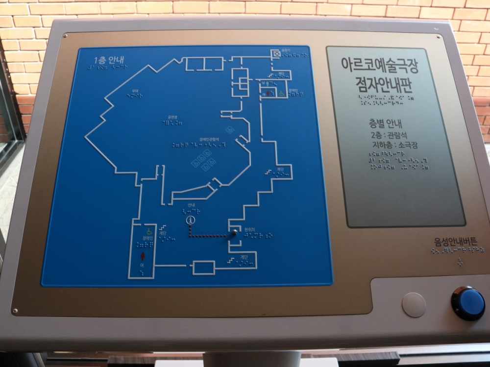 사진 설명 시작. 아르코예술극장 입구의 촉지도이다. 푸른 배경 위에 흰색으로 건물의 경계선이 도톰하게 그려져 있다. 촉지도 아래에는 음성해설을 제공하는 음성안내버튼이 있다. 사진 설명 끝.