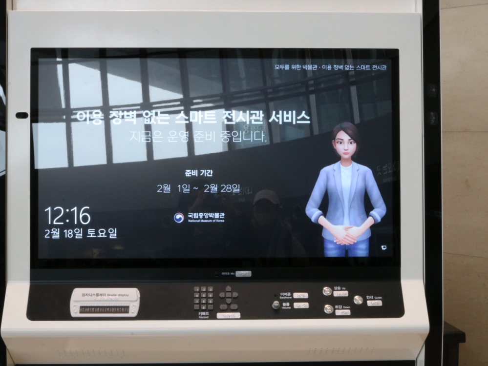 사진 설명 시작. 여러 버튼이 설치된 키오스크 화면이 켜져 있다. 화면에는 다음과 같은 글이 쓰여 있다. 이용 장벽 없는 스마트 전시관 서비스, 지금은 운영 준비 중입니다. 화면 오른쪽에는 손을 모은 여성 안내원의 이미지가 있다. 사진 설명 끝.