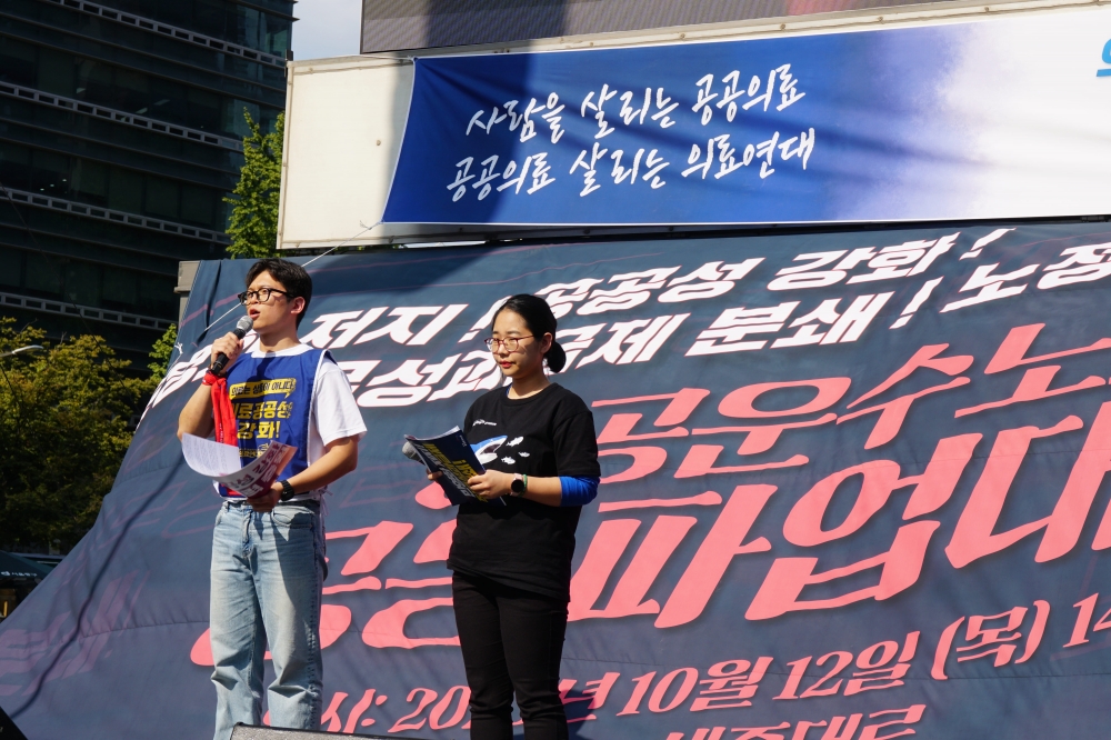 사진설명 시작. 파업 무대 위의 모습이다. 무대 위에는 의료공공성 강화라고 적혀 있는 파란색 옷을 입은 남성과 검은색 옷을 입은 여성이 서 있다. 왼쪽 남자가 마이크를 들고 발언하고 있다. 무대 뒤 현수막에는 빨간 글씨로 공공운수노조 공동파업대회라 적혀 있다. 사진설명 끝.