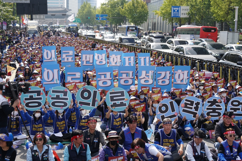 사진설명 시작. 거리에 수많은 사람들이 앉아 있다. 사람들은 의료공공성 강화, 인력충원 등이 쓰인 파란색 옷을 입고 있다. 의료민영화 저지, 의료공공성 강화, 공공의료 인력충원이라 쓰인 큰 피켓을 들고 있는 사람도 있다. 사진설명 끝.