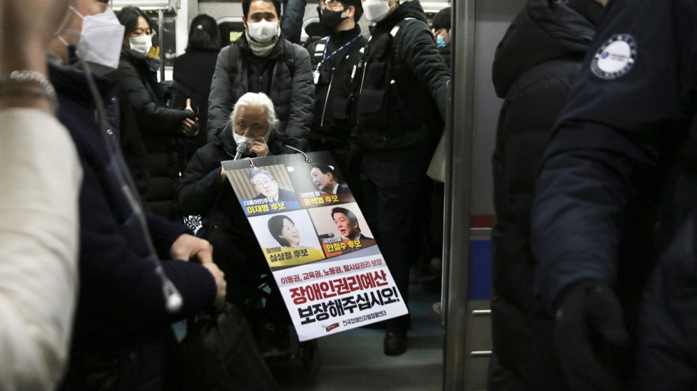 사진 설명 시작. 전국장애인차별철폐연대 회원이 지하철에서 장애인권리예산 보장 시위를 하고 있다. 시위자는 휠체어를 타고 지하철에 탑승하고 있다. 목에는 4명의 대선 후보 얼굴과