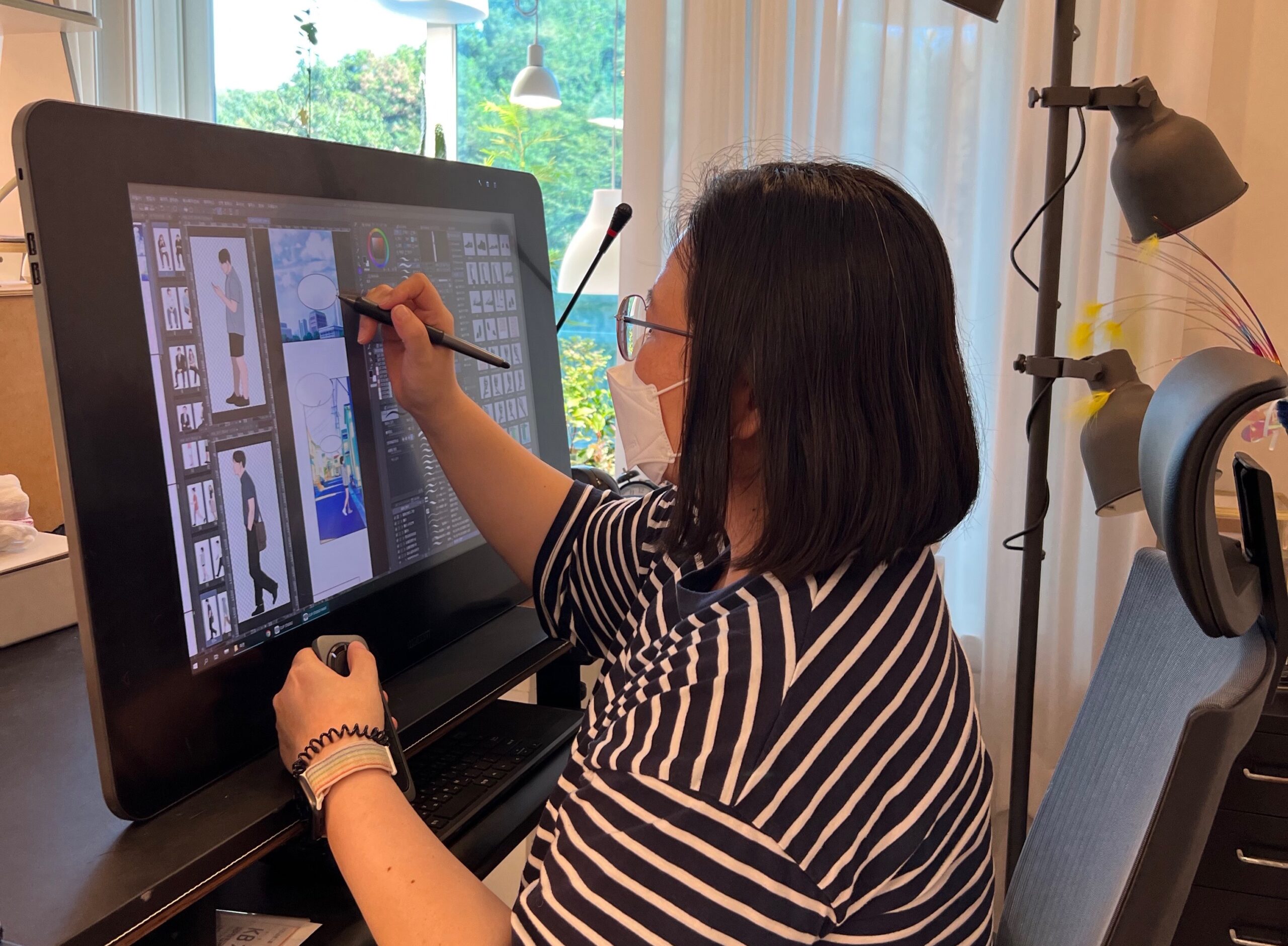 사진 설명 시작. 단발머리, 안경을 쓰고 마스크를 착용하고 검은색과 흰색 줄무늬 반팔 티셔츠를 입은 작가가 의자에 앉아 모니터 화면에 펜을 가져다대고 있다. 화면에는 여러 장의 사람 그림과 풍경 및 말풍선 등 웹툰 작업창이 떠있다. 사진 설명 끝.