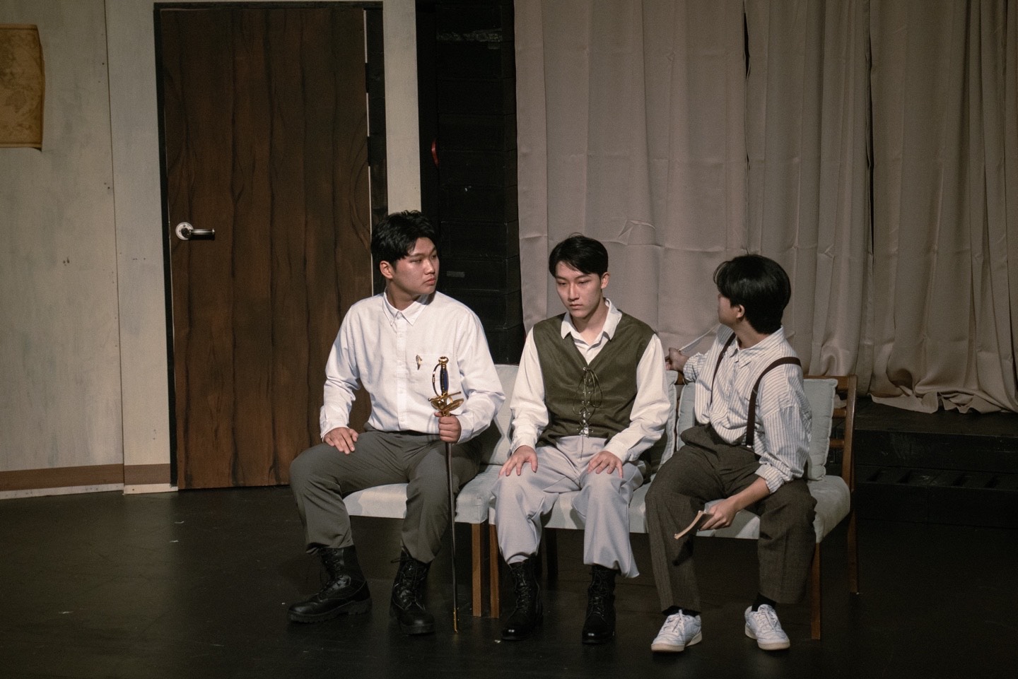 사진 설명 시작. 연극 무대, 세 명의 배우들이 의자에 나란히 앉아 있다. 사진 설명 끝.