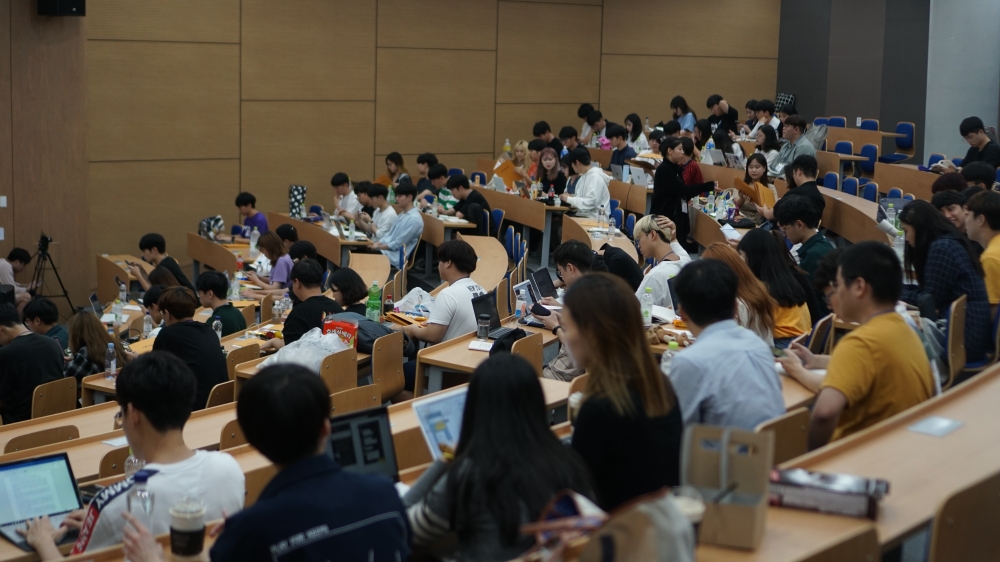 사진 설명 시작. 큰 강의실에 학생들이 앉아 있다. 일부 학생들은 서로 대화를 나누고 있다. 사진 설명 끝.