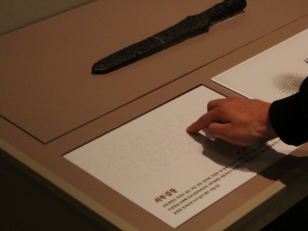 사진 설명 시작. 사진 위쪽에는 고대 시대의 단검이 만질 수 있도록 모형으로 재현된 촉각전시품이 있다. 단검 모형 바로 밑에는 점자와 묵자로 된 유물 설명이 있다. 기자의 손이 점자를 짚어가고 있다. 사진 설명 끝.