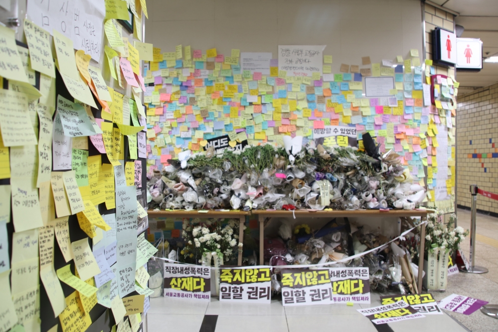 사진 설명 시작. 신당역 여자화장실 앞에 피해자를 위해 추모의 공간이 마련돼 있다. 벽에는 피해자를 추모하는 포스트잇이 붙여있고 책상에는 조화가 놓여있다. 사건의 책임을 묻는 팻말도 놓여있다. 사진 설명 끝.