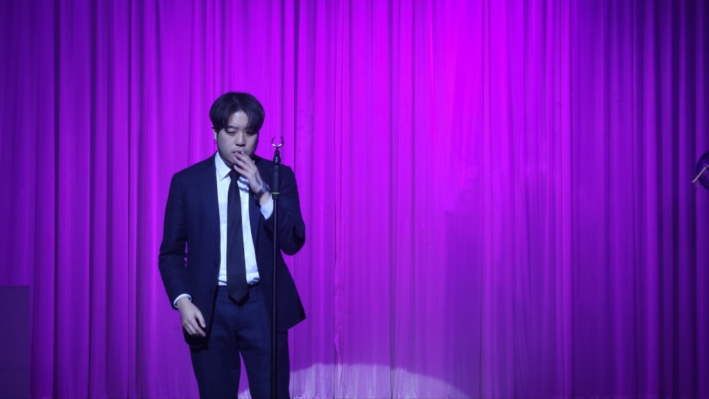 사진 설명 시작. 아무것도 없는 무대 위에 남성 A가 걸어나오고 있다. 앞에는 마이크 스탠드가 덩그러니 놓여 있다. A는 무언가를 생각 중인 듯, 땅을 응시하며 걸어나오는 중이다. 사진 설명 끝.