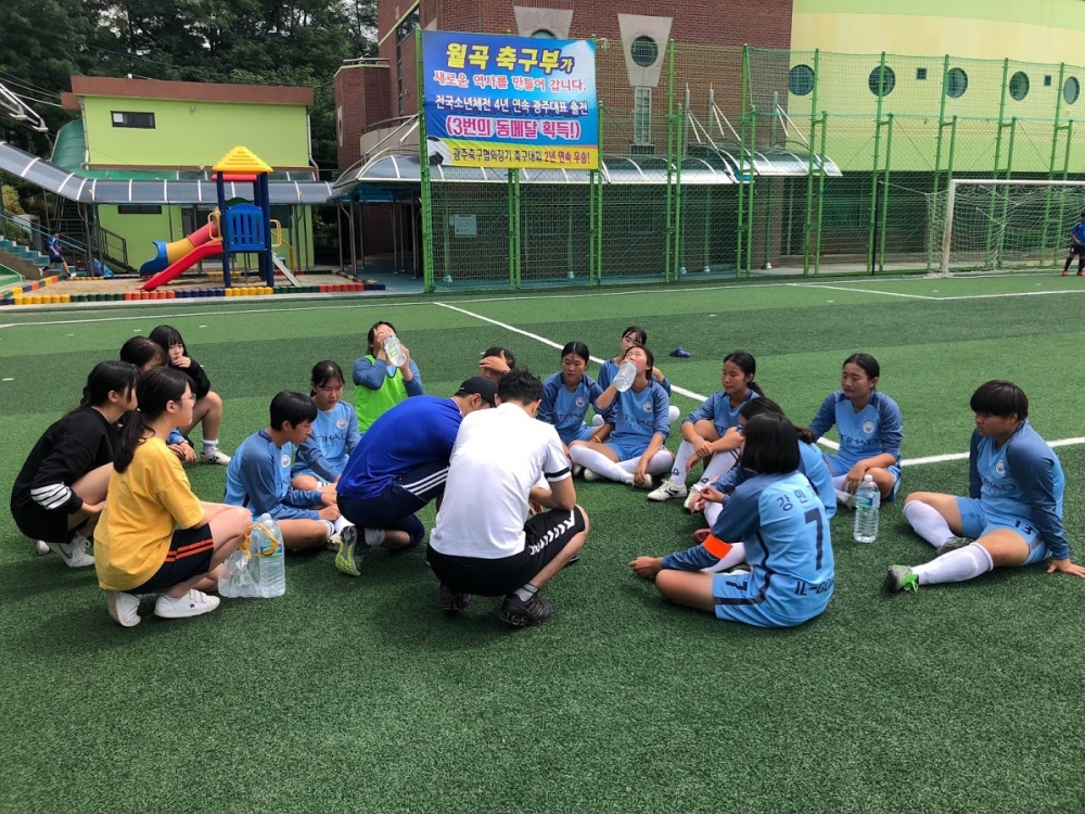사진 설명 시작. 광주 일곡중학교 여자 축구부 학생들이 잔디 위에 앉아 휴식을 취하고 있다. 가운데에는 지도 선생님으로 보이는 남성 두 명이 있고 그 주위에 파란색 유니폼을 입은 열댓명의 여학생들이 벌건 얼굴을 하고 앉아 있다. 그 중 가운데의 두 학생은 생수병을 들고 급히 물을 마시고 있다. 사진 설명 끝.