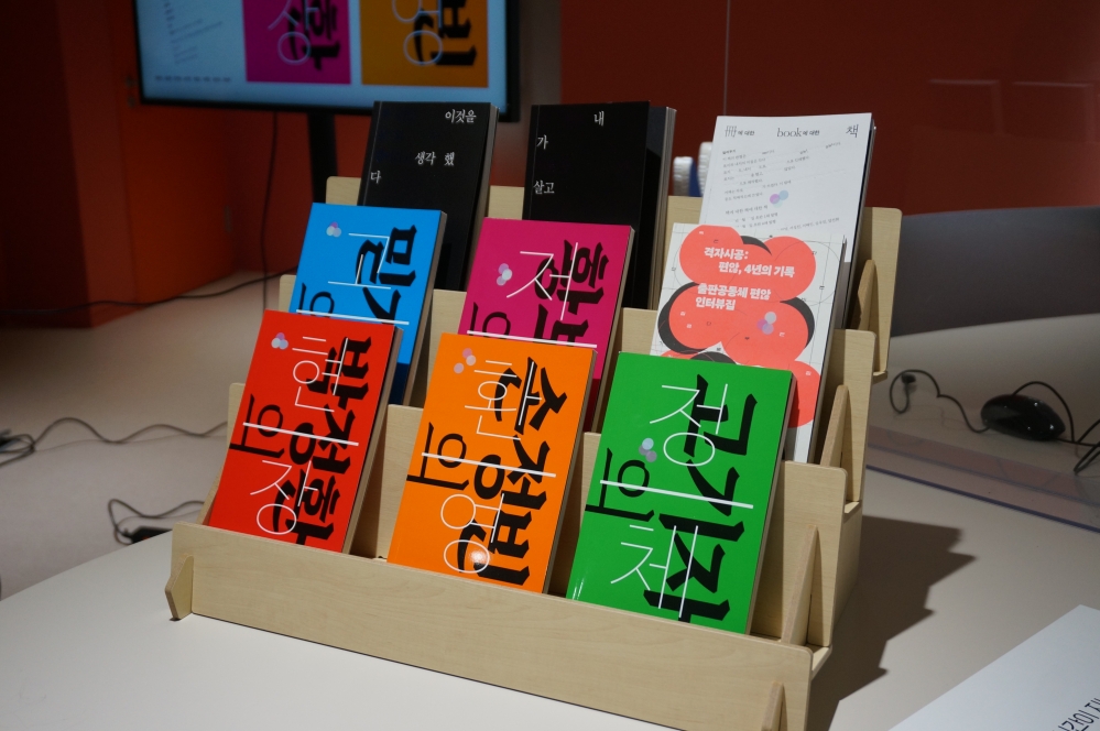 사진 설명 시작. 나무 재질의 선반에 여러 책이 전시돼 있다. 한 줄에 세 권씩 세 줄로 전시되어 있는데, 왼쪽 가장 위부터 검은색 표지의 책 두 권, 흰색 책 한 권이 전시돼있다. 그 아랫줄에는 파란색, 자주색, 코랄색 책이 놓여 있다. 가장 아랫 줄에는 빨간색 표지의