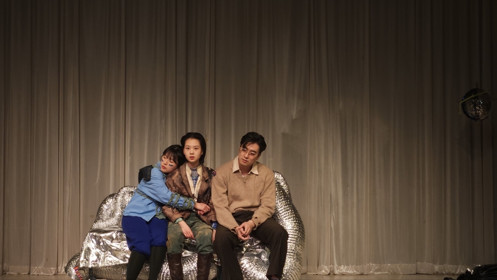 사진 설명 시작. 커튼이 쳐있어 간단해 보이는 배경 앞에 은빛 소파가 놓여 있다. 그 소파에 나란히 앉아 있는 오이디, 이오, 칵터. 극 초반, 오이디 일행이 막막한 일상에서 권태를 느끼고 있는 장면이다. 칵터는 몸이 약해 힘없이 앉아 있고, 오이디는 이오를 꼭 안고 눈을 감고 있다. 이오는 이들을 이끄는 대안 어머니로서 흐트러짐 없이 앞을 응시하고 있다. 사진 설명 끝.