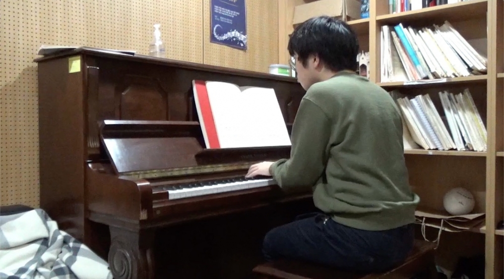 사진 설명 시작. 연습실처럼 보이는 공간에서 한 남자가 피아노를 치는 뒷모습이 보인다. 사진 설명 끝.