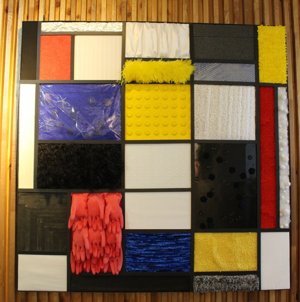 사진 설명 시작. 몬드리안의 그림의 촉각명화가 확대돼 있다. 다양한 크기와 색의 직사각형들로 이루어진 커다란 정방형의 그림이다. 각 직사각형은 해당 색에 어울리는 재질들로 채워져 있다. 예컨대 붉은 정사각형은 빨간 고무장갑들로, 파란 직사각형은 시원한 느낌을 주는 비즈로 채워져 있다. 사진 설명 끝.