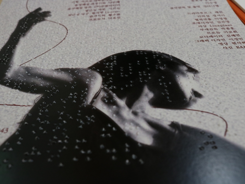 사진 설명 시작. 점자와 묵자가 함께 쓰인 프로그램북을 확대한 사진이다. 회색빛이 도는 종이에 한쪽 팔을 뻗은 여자 무용수의 사진이 흑백으로 삽입돼 있으며, 오른쪽에는 제작진의 직책과 이름이 붉은 글씨로 적혀 있다. 사진 설명 끝.